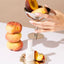 Hand holding a metallic bowl with peaches, a bottle of nail polish, and stacked peaches on a light surface.